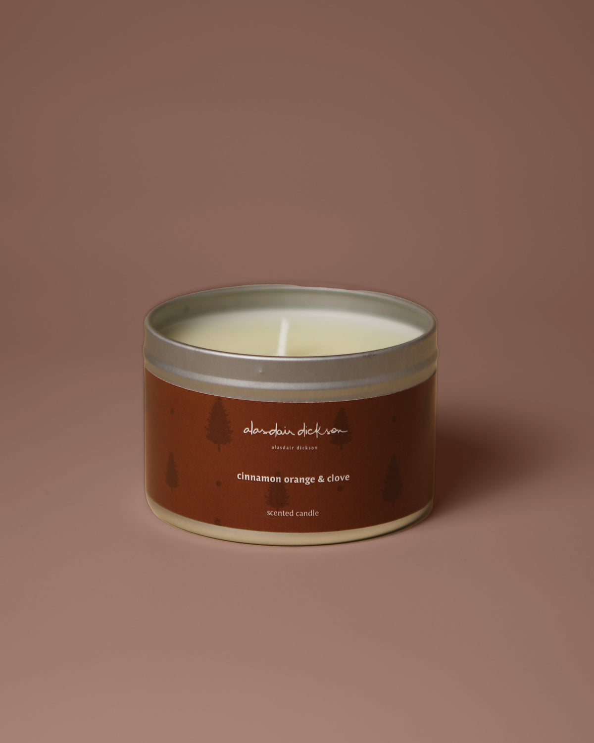 A round metal tin candle with a brown label and silver lid, centered on a solid dusty rose background. The label reads 'alasdair dickson' and 'cinnamon orange & clove'.
