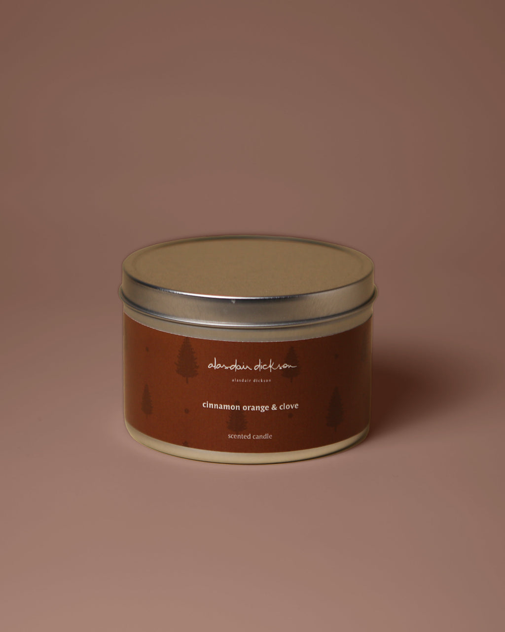 A round metal tin candle with a brown label and silver lid, centered on a solid dusty rose background. The label reads 'alasdair dickson' and 'cinnamon orange & clove'.
