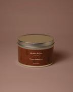 A round metal tin candle with a brown label and silver lid, centered on a solid dusty rose background. The label reads 'alasdair dickson' and 'cinnamon orange & clove'.